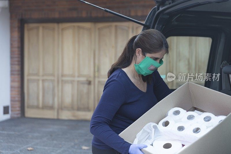 妇女戴着透明面罩和防护手套从汽车后备箱中取出装有食品杂货的盒子。新常态。