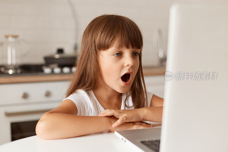 黑头发的小女孩坐在餐桌旁，张着大嘴看着笔记本电脑的显示屏，在厨房里摆造型，惊呆了。