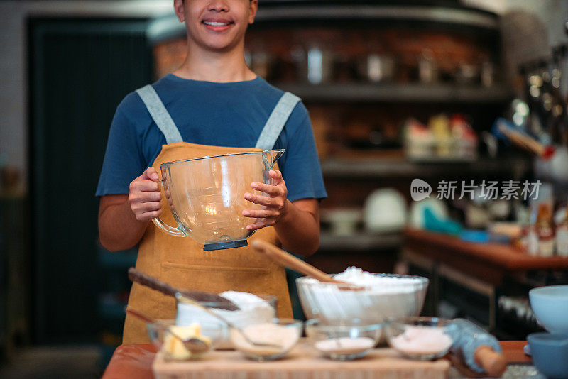 亚洲华人微笑着面包师，十几岁的男孩拿着玻璃量杯在厨房里