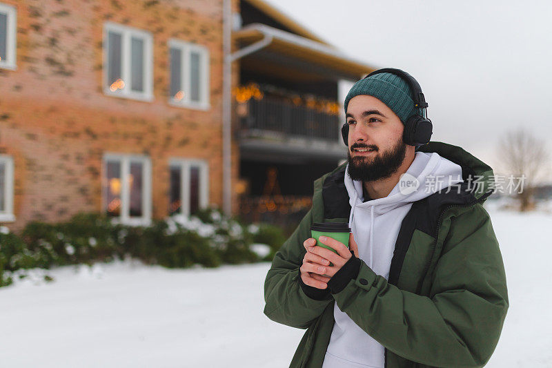 戴着耳机的大胡子青年在室外用一杯热咖啡暖手