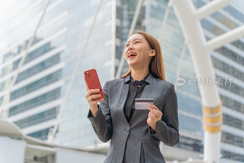 一名年轻的亚洲女商人穿着灰色的办公套装，在商业区用她的智能手机使用信用卡进行在线交易，她微笑着自信