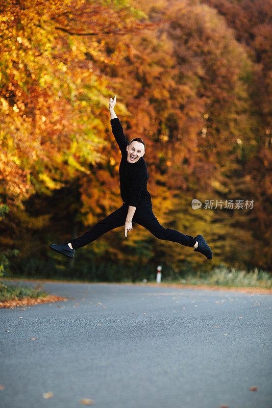 年轻有魅力的男子在秋天的森林路上玩。一个穿黑色衣服的短发男孩在黄叶树的公园里跳跃。乡间惬意的一天