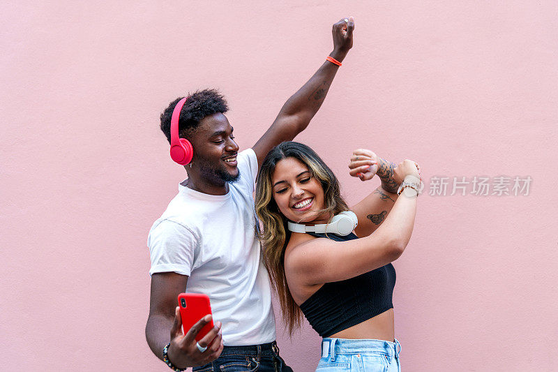 快乐的多元文化组合随着音乐起舞，对着粉红色的墙微笑，都市休闲生活写真。
