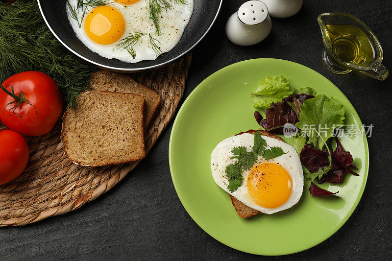 煎锅和盘子上有美味的煎蛋，沙拉和产品放在黑色的桌子上，平铺