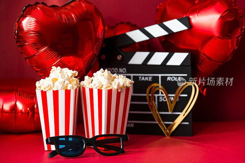 电影工业的一面旗帜。一次浪漫的电影约会。电影摄影机、3D眼镜、爆米花和红色背景上的心形铝箔气球。这部电影在情人节首映。