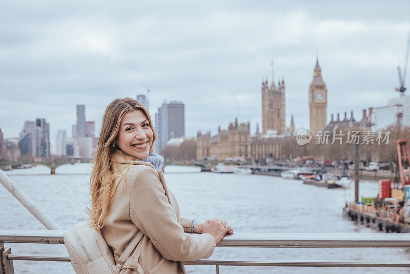 一名30多岁的年轻女子在大本钟附近的金禧桥上摆姿势，面带微笑，对着镜头露出露齿的微笑，似乎在首都伦敦玩得很开心