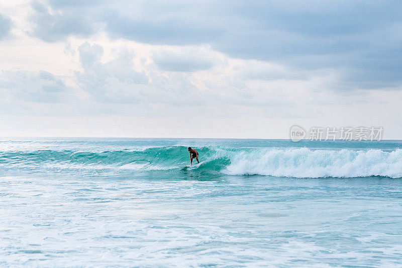 冲浪者骑着绿松石浪:一个熟练的冲浪者在清澈的蓝色海洋中的白色冲浪板上的动态动作镜头。摄于巴厘岛梦幻海滩。