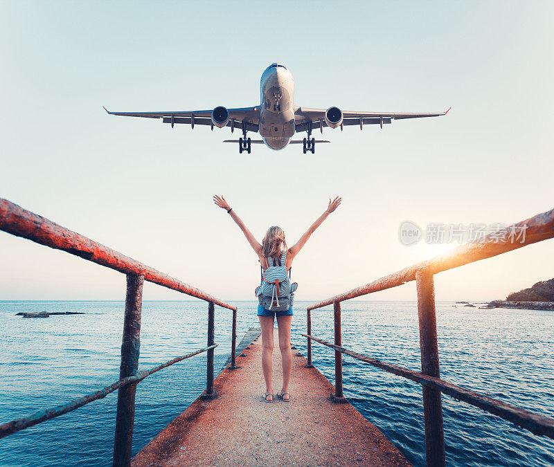 夕阳下的飞机和女人。夏天的风景与女孩站在海上码头举起手臂和飞行的客机。晚上，一名妇女正在降落一架商用飞机。生活方式