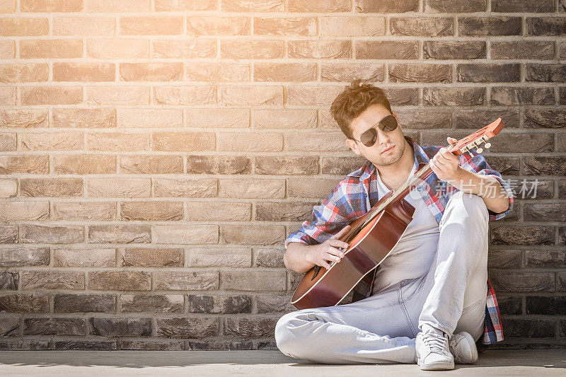 时尚的原声吉他手坐在地上，靠在外面的砖墙上