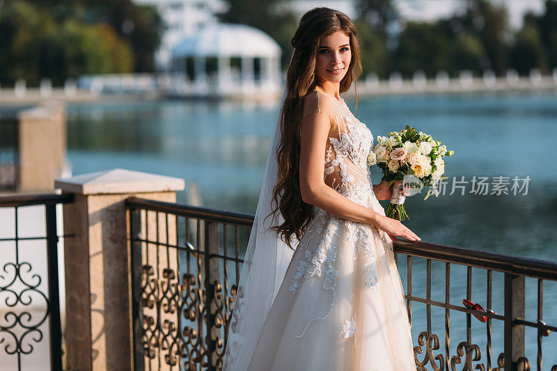 年轻新娘的肖像与长头发拿着她的婚礼花束包括白玫瑰和其他鲜花。漂亮的白色婚纱。美丽的女孩在湖的背景