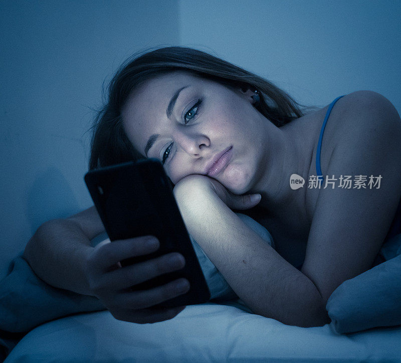 年轻漂亮的女人沉迷于床上，用她的智能手机聊天和上网困倦，无聊和疲惫的深夜在移动上瘾和技术过度使用的概念。