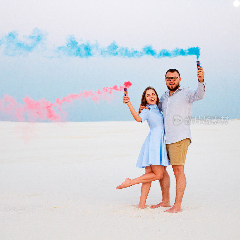 年轻的情侣站在沙滩上，手里拿着彩色烟雾弹，浪漫的情侣带着蓝色和红色的烟雾弹在沙滩上