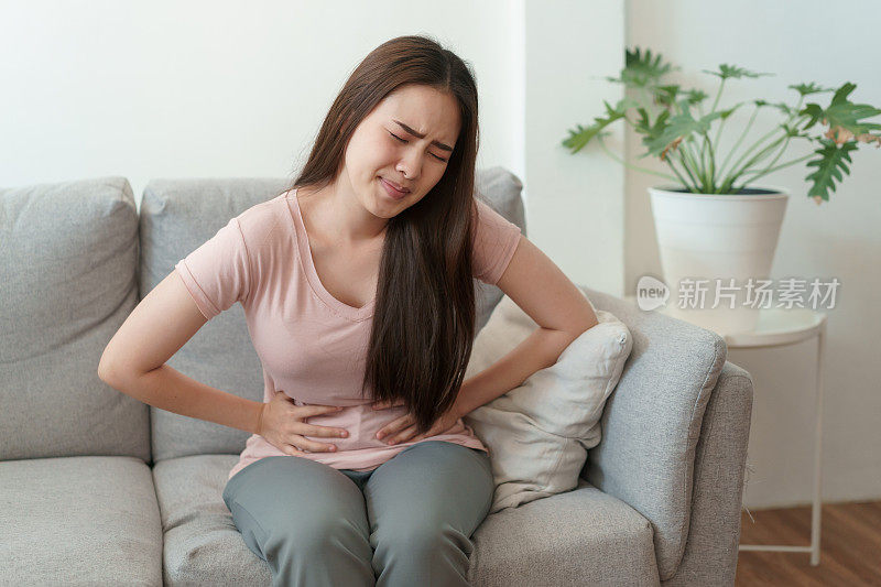 有月经和痛经的亚洲妇女。年轻女性坐在她家里的沙发上痛苦