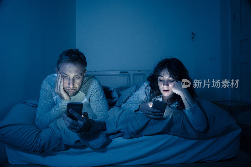 这是一对无聊的年轻情侣晚上躺在床上玩手机，沉迷于游戏、社交媒体和应用程序，忽视彼此的生活方式。关系沟通问题和手机成瘾的概念。