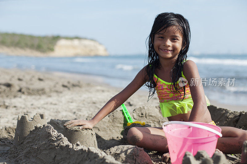拉丁裔女孩坐在沙上用桶和铲子建沙堡。