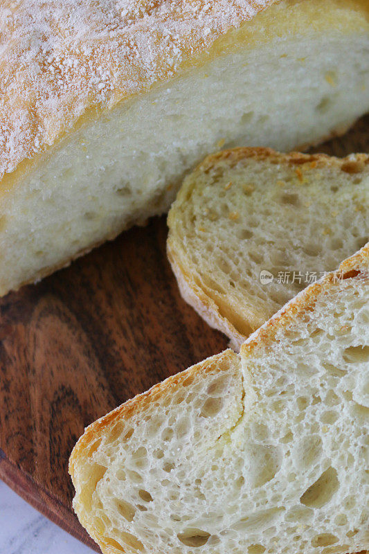 这是自制的查巴塔面包的特写，放在圆形的木制砧板上，金黄色，刚出炉的面包，硬壳的面包被切成薄片，从高处俯瞰