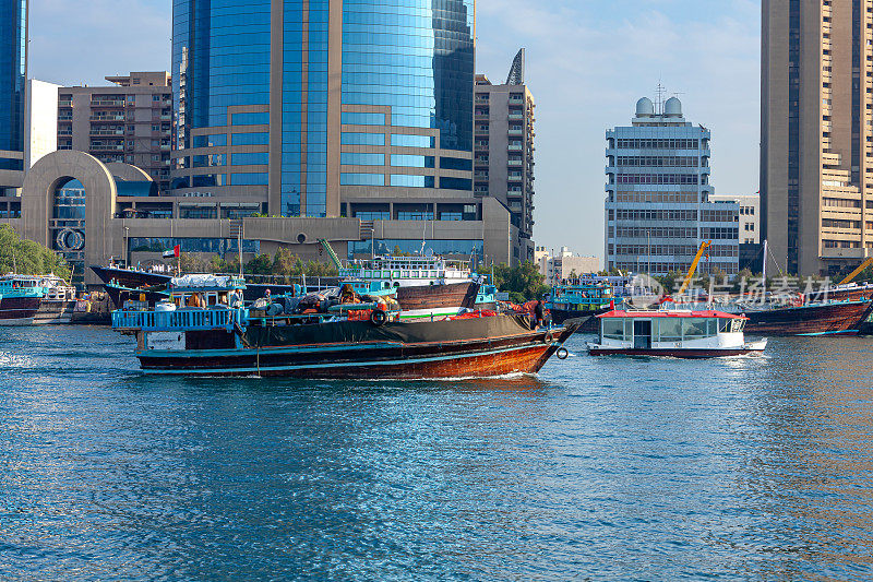阿拉伯联合酋长国迪拜——晨曦中，一艘满载的传统阿拉伯单桅帆船驶入迪拜河——背景中，在德拉一侧，单桅帆船彼此并排停泊。在遥远的背景，办公室和住宅建筑