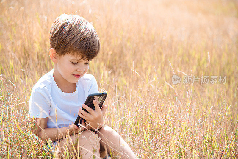 夏天日落时，兴高采烈的孩子坐在草地上，看起来就像手机里的卡通。可爱的男孩在大自然中玩耍
