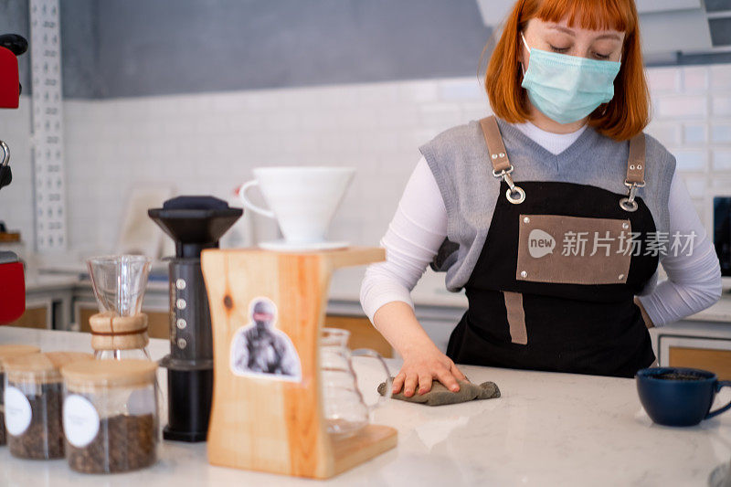 年轻的女咖啡师戴着口罩和清洁咖啡店吧台