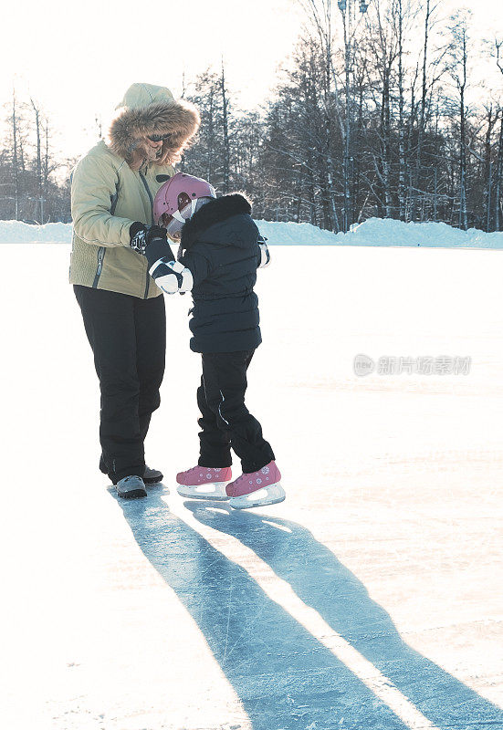 教孩子溜冰。一个年轻的女人在城市的室外溜冰场训练一个小女孩。