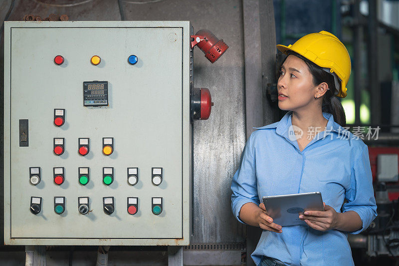 亚洲女性工业工厂工人，戴着黄色安全帽，手拿平板电脑，站在工厂的电子机器控制面板旁工作。工业工厂工人日常工作的概念