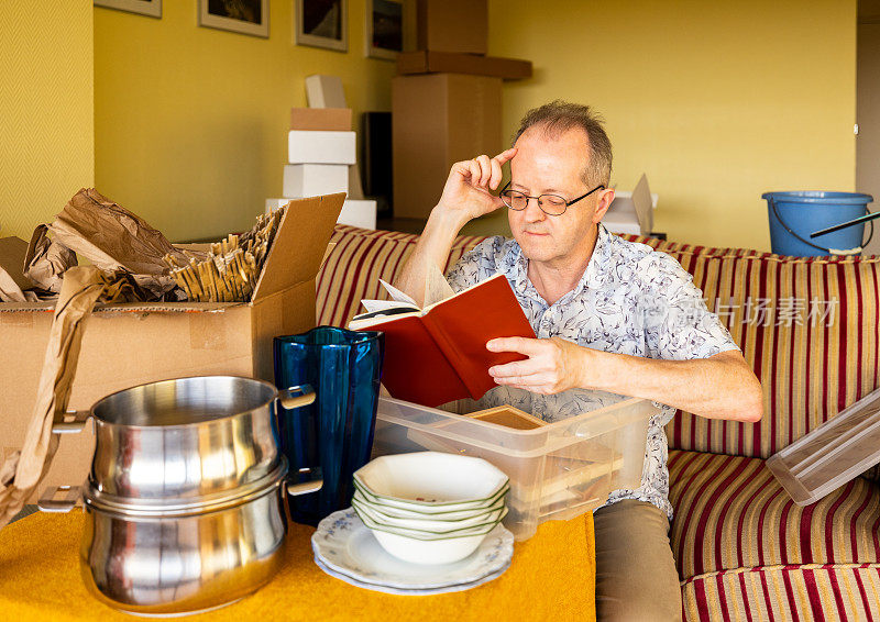 一个活跃的老人独自坐在沙发上读一本旧书，同时打开纸箱搬家或公寓