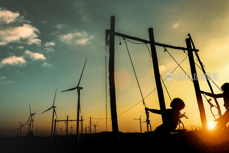 孩子们幸福生活的剪影，在夕阳下可再生的风力涡轮机公园玩秋千。企业社会责任和可持续能源概念。电影的暖色。