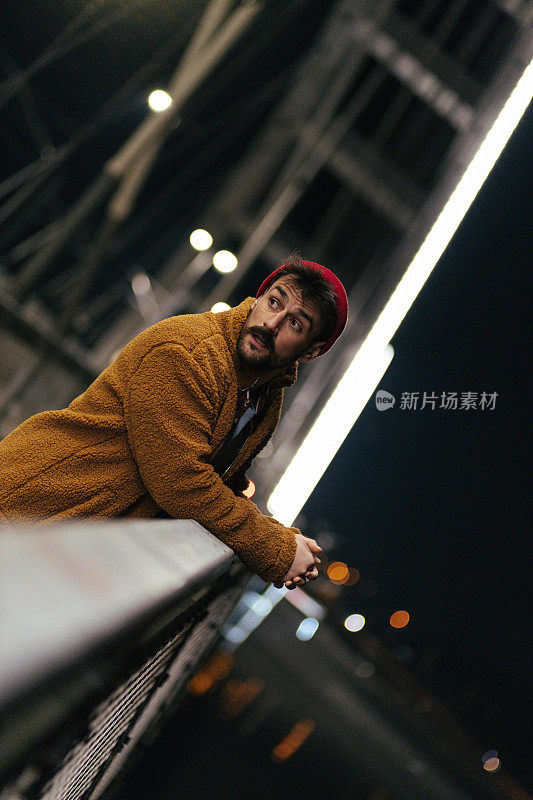 一个年轻英俊的时髦男人的肖像与胡子倚靠在桥栏杆在晚上