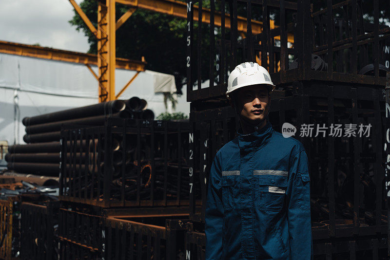 金属工业-匿名肖像的工人戴着安全帽的眼睛覆盖在深的阴影前站在一个开放的仓库装满了球墨铸铁管和零件