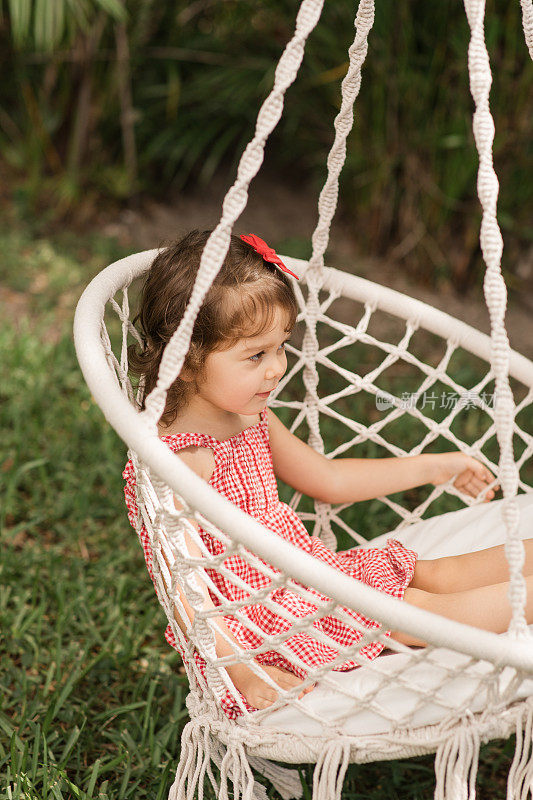 一个3岁的蹒跚学步的女孩放松在一个波西米亚流苏户外秋千挂在南佛罗里达州的一棵大树上，一个节日和爱国的美国庆祝野餐在炎热的夏天