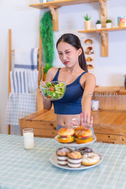 照片年轻美丽的亚洲女性女士在运动服装说不和拒绝诱惑汉堡和甜甜圈在厨房的桌子上，选择一碗沙拉与樱桃番茄和一杯牛奶的饮食和减肥健康的生活方式