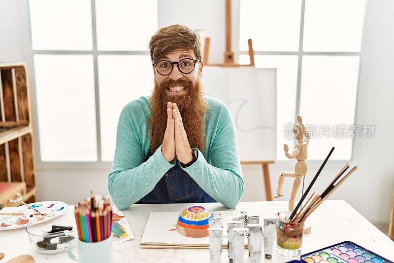 红发长胡子男子在艺术工作室画泥碗，双手合十祈祷，请求宽恕，微笑自信。
