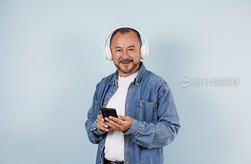 拉丁美洲拉丁美洲高级男子手持手机听音乐，耳机在蓝色背景上的复制空间