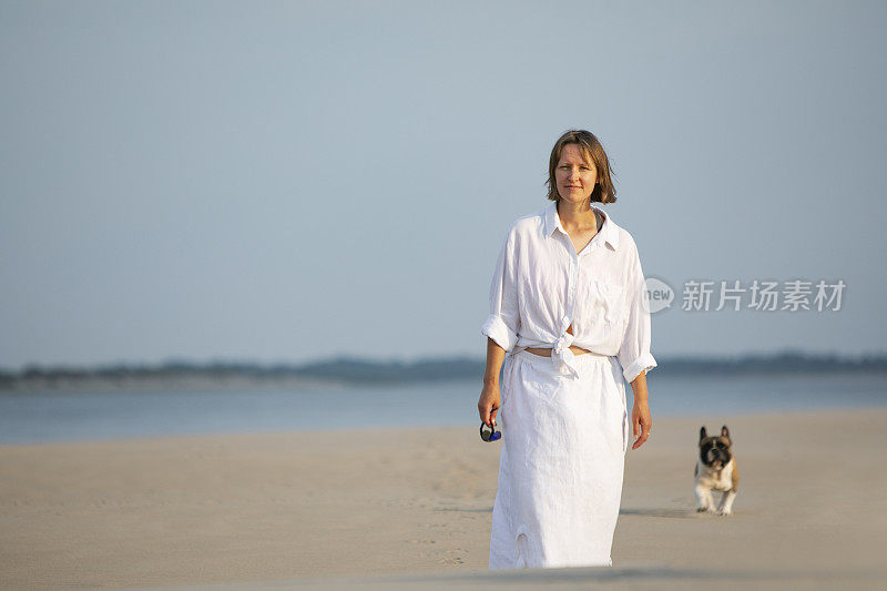 年轻的成年女子和她的法国斗牛犬在海滩上散步