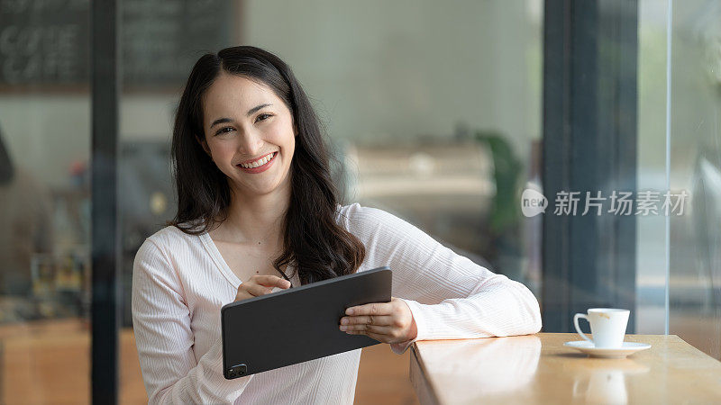 年轻快乐的亚洲女子手持平板电脑，站在咖啡馆里对着相机微笑的肖像。