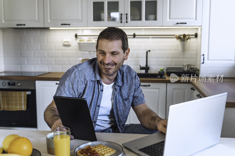 一名男子早上在厨房餐桌边吃早餐时使用笔记本电脑。