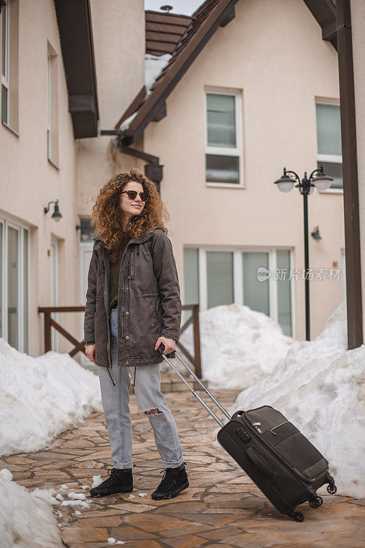 一位年轻的女旅行者提着行李箱站在一家旅馆门前，她是来度寒假的