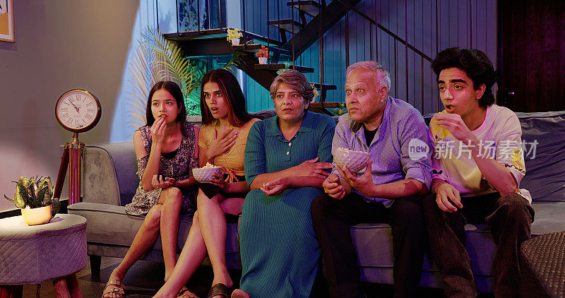 印度幸福延伸的老年家庭坐在沙发上看恐怖惊悚电视，脸上带着恐惧的表情，年长的祖父母，年轻的十几岁的男孩，女孩，拿着碗，一起吃零食爆米花，深夜在家里看电影