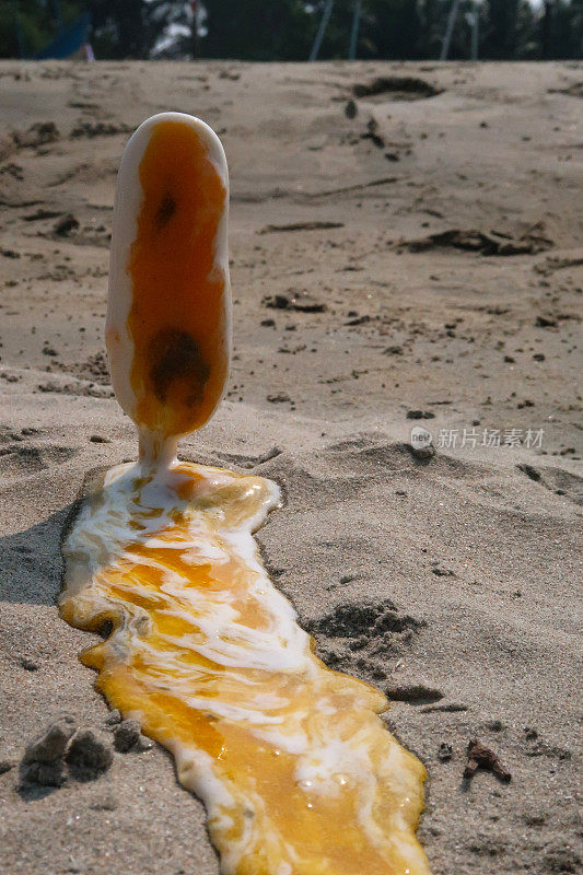 图像融化的冰淇淋棒棒糖与木棒棒糖棒卡在沙滩上的沙子，橙色的冰棒在退潮时在海水的边缘，芒果和橙色泥覆盖的冰淇淋pop反对海岸背景，重点在前景