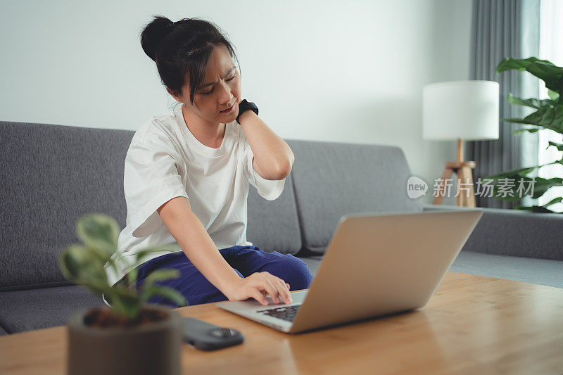 一名亚洲女性坐在家里的沙发上使用笔记本电脑，身体疼痛。