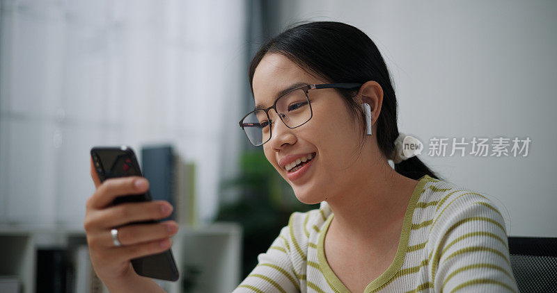 年轻的亚洲女性戴着眼镜和耳机，坐在办公桌上用手机视频通话