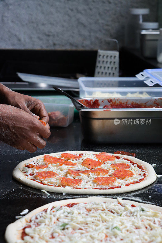 面目全非的Pizzaiolo(披萨厨师)在未煮熟的披萨基础上添加意大利辣香肠片，意大利辣香肠披萨富含番茄马里纳拉酱，磨碎的马苏里拉奶酪，商业厨房设置，重点放在前景