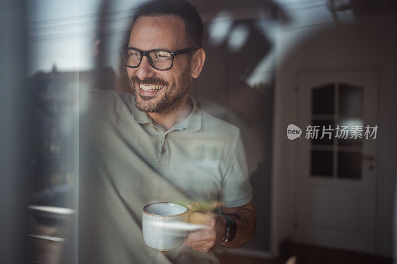 一位成年男子端着一杯咖啡，面带微笑地站在公寓的阳台门口