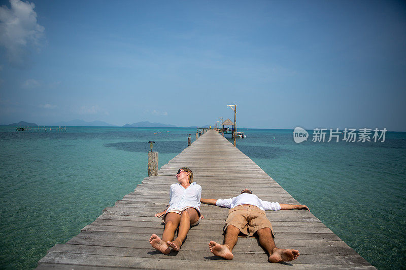 一对年轻夫妇躺在热带岛屿上的木码头上