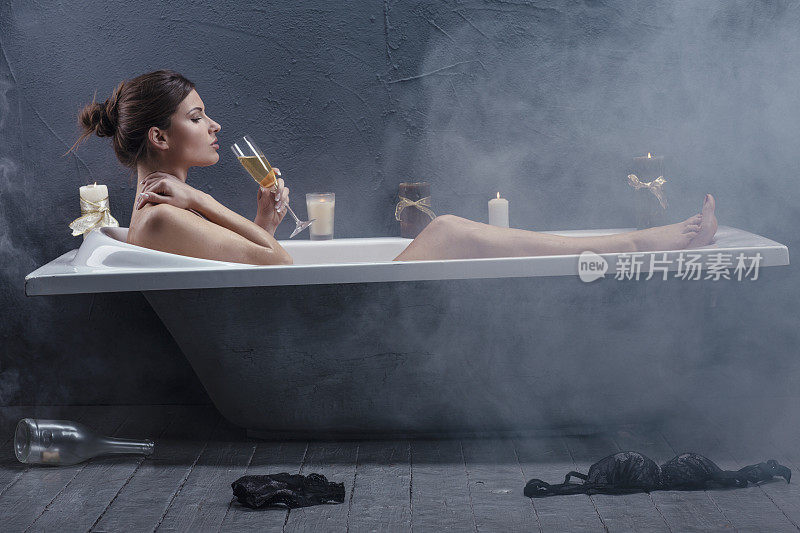 美丽的女人在浴缸里放松，点燃蜡烛。29岁的女人在洗澡喝香槟。潮湿的。新时代美容与健康