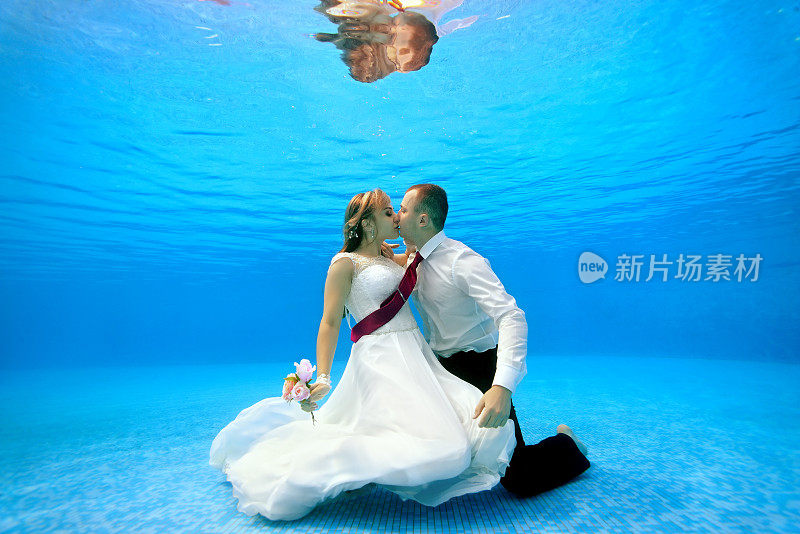 穿着婚纱的新娘和新郎在游泳池底部拥抱亲吻。肖像。横向。在水下拍摄