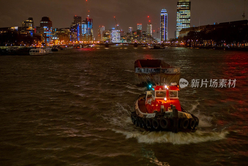 夜晚的泰晤士河，以伦敦西区的天际线为背景，一艘拖船拖着一艘空驳船。