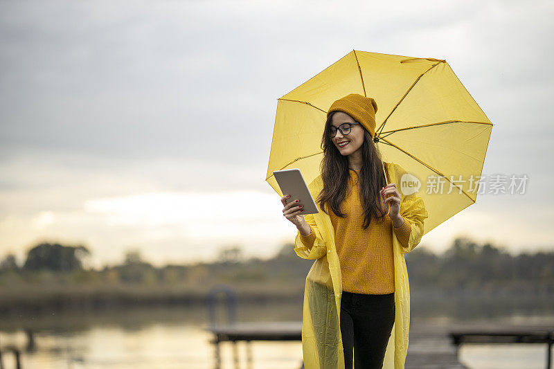 女人用着平板电脑，撑着伞，走在湖边的码头上