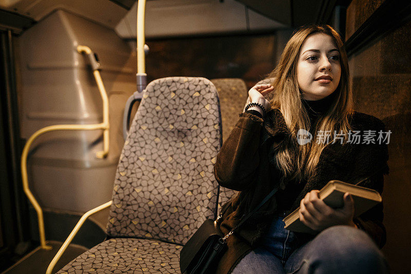 晚上乘公交车时拿着书的女乘客。晚上，女孩在窗口旁边的移动车辆上旅行