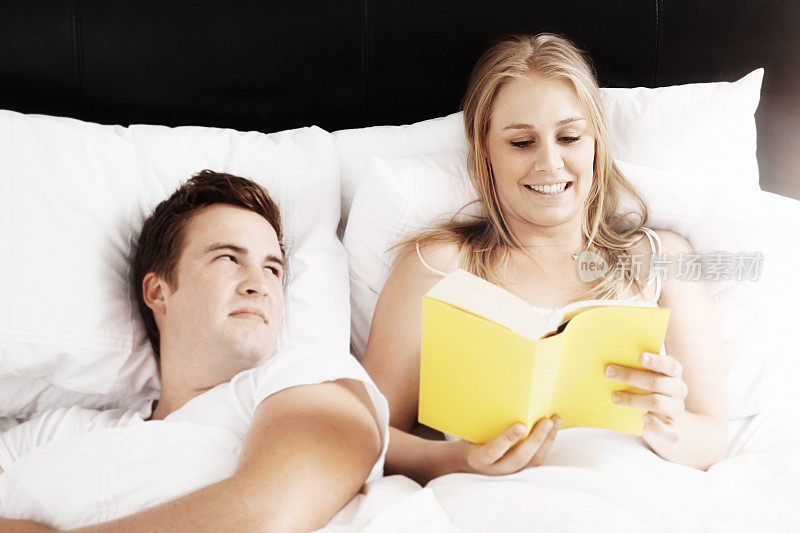 一对夫妇躺在床上:男人给女人一个充满爱意的眼神，而她正在看一本平装书